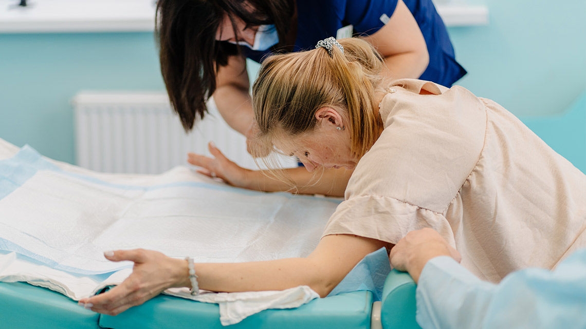 Una donna in un ambiente sanitario, inclinata in avanti su una superficie imbottita coperta da materiale monouso, con le braccia protese per sostenersi. Un operatore sanitario con una maglia scura e una mascherina assiste nelle vicinanze, mentre il braccio di un’altra persona offre ulteriore supporto. L’ambiente clinico interno presenta una parete chiara e un termosifone, suggerendo un momento di sforzo fisico, probabilmente durante il travaglio.