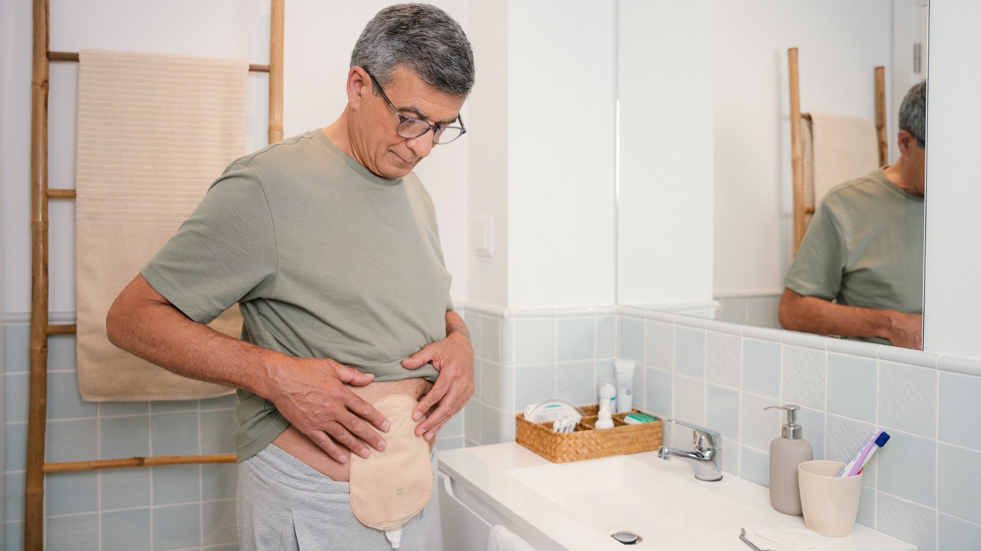 Un uomo che indossa gli occhiali e una maglietta verde guarda verso il basso la sua sacca per urostomia, fissata all'addome, mentre si trova in piedi in un bagno. Accanto a lui è visibile il suo riflesso in uno specchio.
