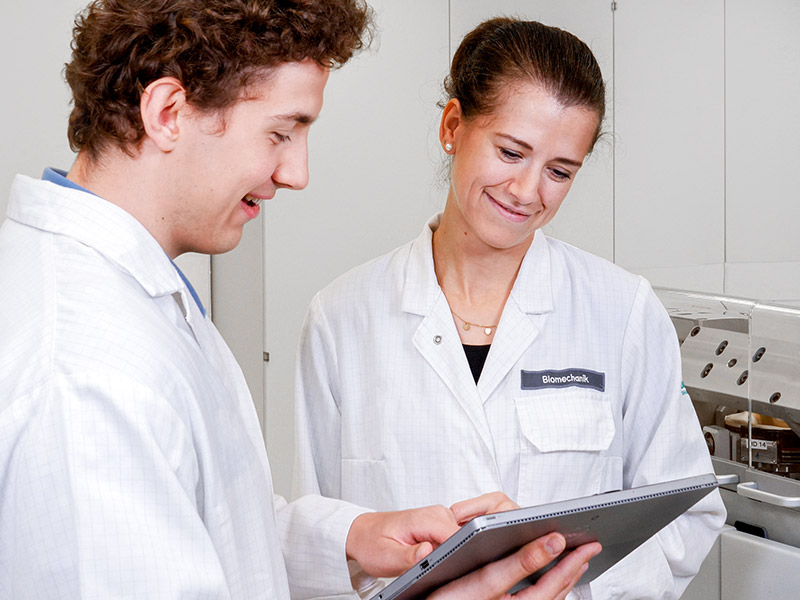 A male healthcare professional is holding a tablet and pointing to something on the screen, showing it to a female healthcare professional